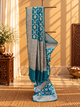 A beautiful, elegant peacock-blue Banarasi-style saree featuring intricate silver zari floral vines and delicate pink floral motifs. The saree has a rich, ornate silver border embellished with pink flowe