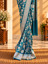 A beautiful, elegant peacock-blue Banarasi-style saree featuring intricate silver zari floral vines and delicate pink floral motifs. The saree has a rich, ornate silver border embellished with pink flowe