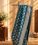 A beautiful, elegant peacock-blue Banarasi-style saree featuring intricate silver zari floral vines and delicate pink floral motifs. The saree has a rich, ornate silver border embellished with pink flowe