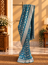 A beautiful, elegant peacock-blue Banarasi-style saree featuring intricate silver zari floral vines and delicate pink floral motifs. The saree has a rich, ornate silver border embellished with pink flowe