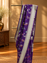 A stunning royal purple saree adorned with delicate multicolour floral and vine motifs, beautifully complemented by a broad silver zari border featuring intricate traditional weaving.