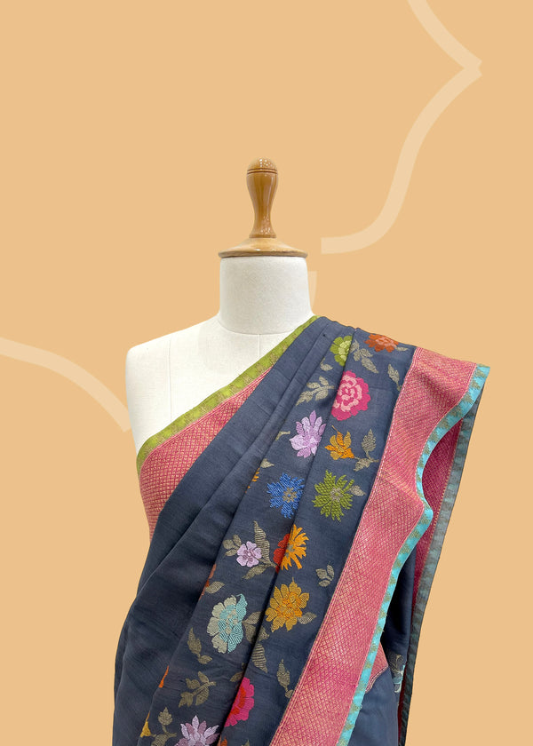 A charcoal gray tussar Georgette saree with multicoloured flowers scattered all over the border and pallu of the saree with a contrast pink woven border and ganga jamuna firozi and olive green kanni.  Shop the best collection of authentic, handwoven, pure benarasi sarees with Roliana New Delhi