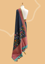 A charcoal gray tussar Georgette saree with multicoloured flowers scattered all over the border and pallu of the saree with a contrast pink woven border and ganga jamuna firozi and olive green kanni.  Shop the best collection of authentic, handwoven, pure benarasi sarees with Roliana New Delhi