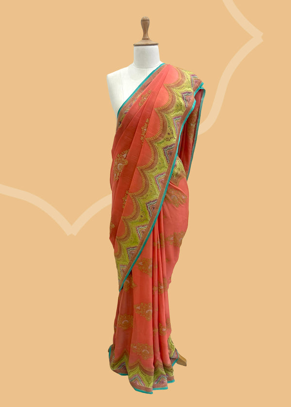 A salmon peach saree in Georgette fabric with a detailed scallop border hand-painted in defferent shades of green,pink firozi with zari ornament bootas and a firozi kanni. A pure Banarasi wedding Sari Shop the best collection of authentic, handwoven, pure benarasi sarees with Roliana New Delhi
