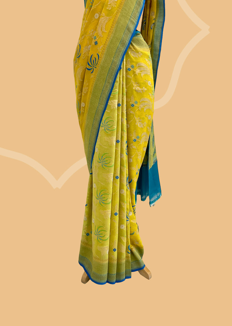 A bright yellow saree in Georgette with contrast blue meenakari floral jaal with zari work coordinated in a simple refreshing design. A pure Banarasi wedding Sari Shop the best collection of authentic, handwoven, pure benarasi sarees with Roliana New Delhi